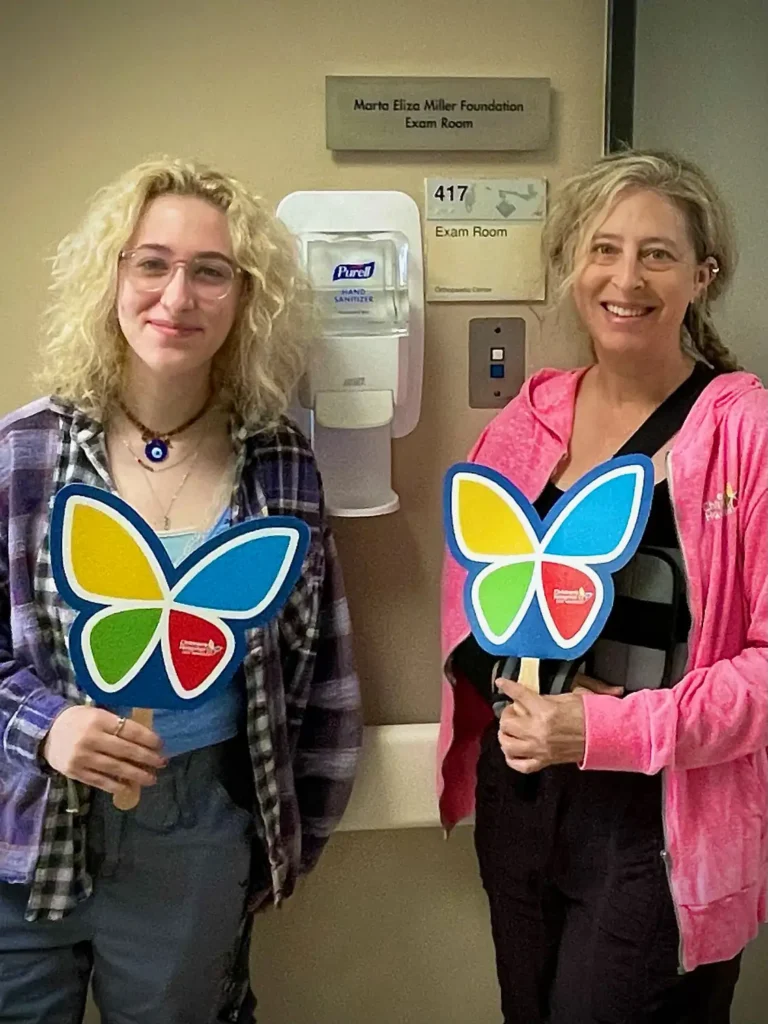 Marta and her mother Corrina stand in front of a plaque outside an Exam room at CHLA that reads Marta Eliza Miller Foundation Exam Room. They both hold the CHLA logo
